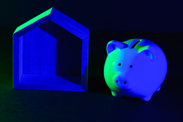 Piggy bank and toy Wooden house on a dark background with green-blue backlight.  The concept of buying a new home, mortgage, renting