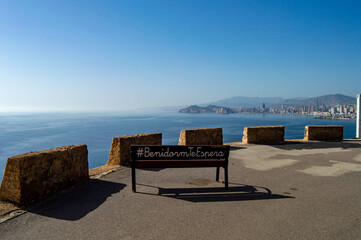 banco de madera en lo alto del mirador de Benidorm con el hashtag Benidorm te espera escrito en castellano mirando hacia toda la ciudad.