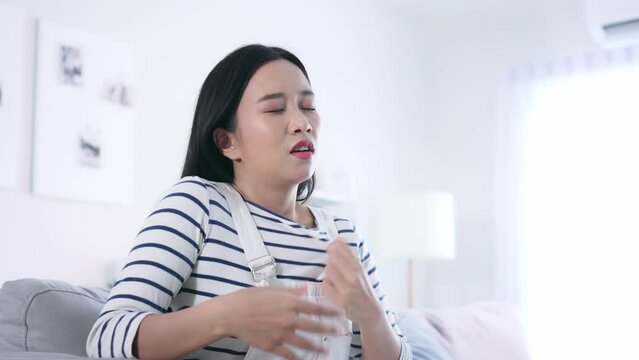 Young unhappy Asian woman suffering the summer heat while sitting on sofa in living room at home. Air conditioner system broken.
