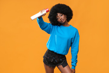 Black-skinned model with an afro hairstyle and spray paint in her hand on an orange background.