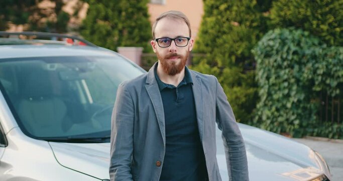 Portrait middle age businessman in glasses standing near parked car in the parking look at camera smiling.