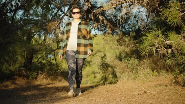 A girl in leggings and a warm plaid shirt walks through a sunny coniferous forest.
