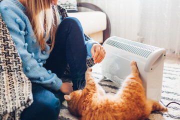 Using heater at home in winter. Woman playing with cat by electric device wearing sweater and hat...