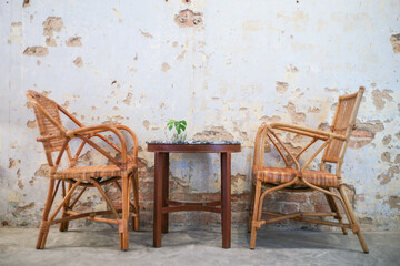 Couple of rattan chairs with the round wooden table in front of the old white bricklayer wall in...