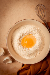 Food background, concept, plate with wheat flour, broken egg, whisk, kitchen towel, on a beige table, top view, rustic,