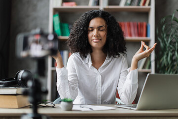 Dark hair curly woman freelancer sitting at table with closed eyes recording blog how to relieving stress by meditation at workplace. Concept of relaxation and harmony, no stress free relief at work.