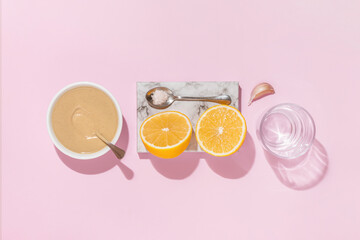 Ingredients for garlic tahini sauce with halved lemon and glass of water on isolated pastel pink background with shadows and reflections. Aesthetic concept of preparing food. Minimal flat lay.