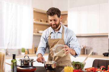 Smiling handsome adult caucasian male chef with beard in apron prepare porridge and salad in modern kitchen - obrazy, fototapety, plakaty