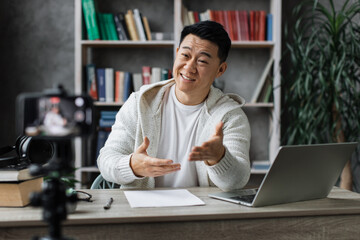 Handsome asian man in casual clothes talking and gesturing while recording video on modern phone. Male blogger sitting at home and doing live stream.