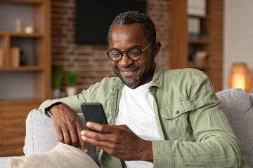 Smiling middle aged african american man in glasses reads message on smartphone, watches video
