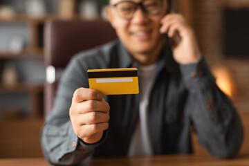 Happy adult asian male in glasses looks at credit card, sit at table, calls by phone in home office interior, cropped