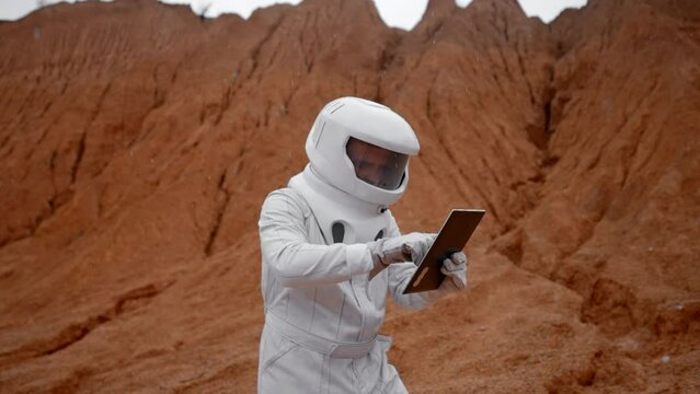 The astronaut is trying to fix a non-working tablet. On the distant planet Mars, an cosmonaut in a white suit and helmet tries to explore the surface using a tablet