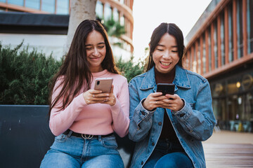 Two girls friends having fun while sitting on a bench and texting their friends on the mobile phone