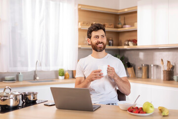 Fototapeta na wymiar Glad adult caucasian guy with beard in white t-shirt enjoys cup of coffee, sits at table with laptop, thinks