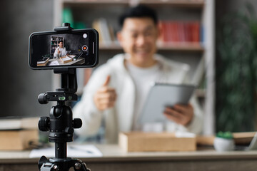 Focus on smart phone screen , asian man recording video on phone camera while unpacking box with new tablet. Young male influencer sharing with subscribers his positive feedback about new order.