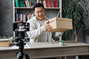 Handsome asian man in casual wear recording video on camera while unpacking gift box. Male blogger sharing his emotions with his subscribers in social networks.