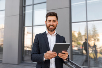 Cheerful confident attractive young european male ceo manager with beard in suit typing on tablet
