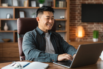 Cheerful attractive mature asian businessman look at computer at table, has video call at home office interior