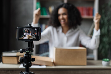 Fototapeta na wymiar Pretty multinational woman in casual wear recording video on camera while unpacking gift box. Female blogger sharing her emotions with her subscribers in social networks.