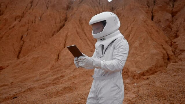 Dolly out of an astronaut working on laptop on an unknown red surface planet. White helmet and special guarding suit for non-atmosphere environment. Slow motion.