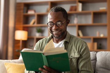 Portrait of cheerful middle aged african american male with glasses reading book in living room...