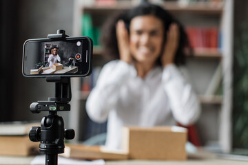 Focus on smart phone screen, woman recording video on phone camera while unpacking box with new wireless headphones. Female influencer sharing with subscribers her positive feedback about new order.