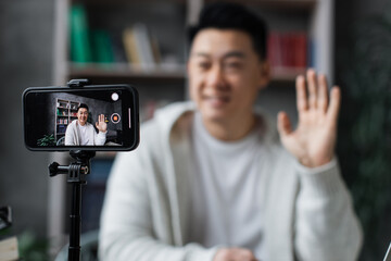 Focus on screen, attractive asian man in casual clothes talking and gesturing while recording video on modern phone on tripod. Male blogger sitting at home and doing live stream.