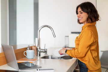 Smiling Arab Woman Washing Dishes And Watching Videos On Laptop In Kitchen