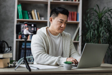 Pleasant young asian man working on laptop while recording video on his smart phone on tripod. Handsome male freelancer working remotely while staying at home.