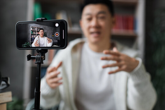 Focus on screen of smart phone, asian man in casual clothes talking and gesturing while recording video on modern phone on tripod. Male blogger sitting at home and doing live stream.