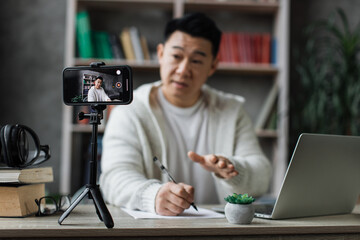 Focus on screen, smiling asian man talking and gesturing in front of modern smartphone camera on tripod, writing report while sitting on background of modern home.