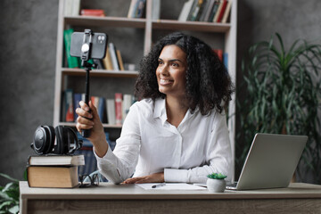 Charming multinational woman in white shirt taking selfie on modern phone on stick. Female blogger...