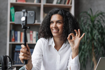 Charming multinational woman in white shirt talking and gesturing while recording video on modern...
