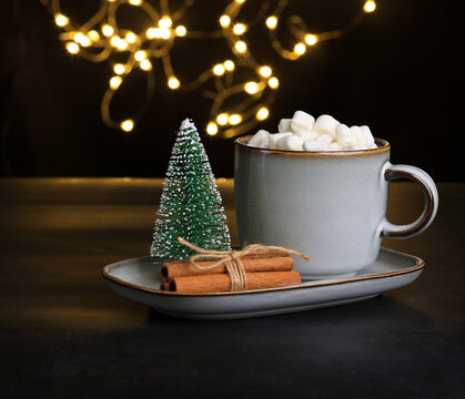 A cup of hot chocolate or cocoa with marshmallow, small Christmas tree and cinnamon sticks on the plate near the cup, defocused Christmas lights in the back