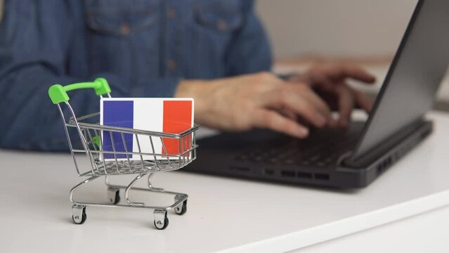 Small shopping cart with flag of France on the table. Man using laptop for shopping online.