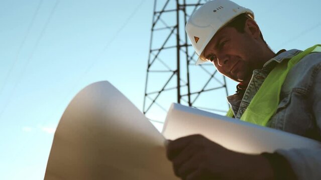 Worker male engineer using documents for checking data while standing against high voltage power towers. Power engineering specialist with working documentation working near electric poles.