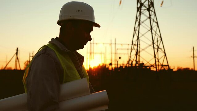 Silhouette of engineer walk on field with electricity towers. Electrical engineer with high voltage electricity pylon at sunset background. Power workers at work concept.