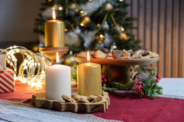 Three burning candles of white and gold color on wooden stands on a red and white tablecloth of the Christmas table