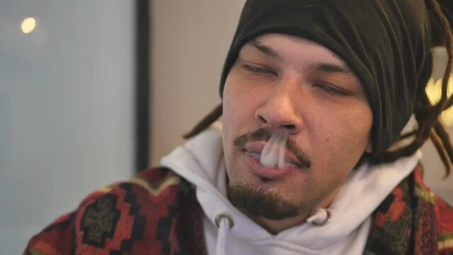 close-up of a young handsome man with mixed race dreadlocks smokes and releases thick tobacco smoke from his mouth into the nostrils of his nose. cute rastafarian lets out cigarette smoke up his nose