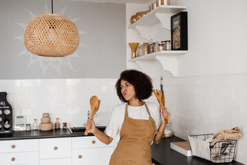 Happy african housewife in apron singing and dancing on the kitchen before cooking breakfast. African american cook girl dancing with spatulas for cooking and having fun.