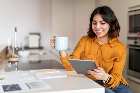 Smiling arab woman drinking coffee and using digital tablet in kitchen