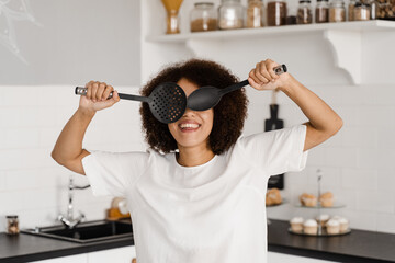 African american girl cook having fun on the kitchen. Advertising for cooking courses and food workshops. African housewife in apron is posing with spatulas for cooking on the kitchen.