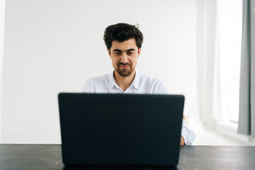 Studio portrait of smiling bearded man in shirt working typing on laptop computer looking to screen sitting at desk in room with light interior. Positive freelancer male having distance work at home.