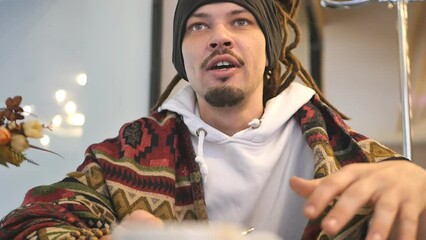An attractive young guy with dreadlocks and a plaid on his shoulders with a charming look and green eyes in a restaurant at a table is happy with the cappuccino he brought, thanks the waiter smiling