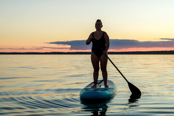 a woman in a closed swimsuit with a mohawk standing on a SUP board with an oar floats on the water against the background of the sunset sky.