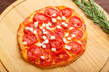 Homemade pizza with tomatoes and mozzarella on a round wooden board next to a sprig of fresh rosemary.