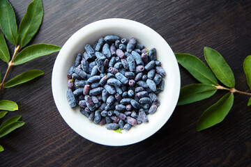 Fresh honeyberry (Lonicera caerulea) in white bowl on wooden table, top view, flat lay. Blue haskap berry with green leaves