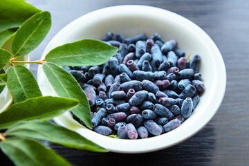 Fresh honeyberry (Lonicera caerulea) in white bowl on wooden table. Blue haskap berry with green leaves