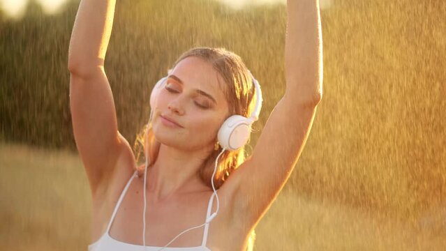 Young beautiful blonde woman in headphones enjoying music under warm summer rain. An incredibly aesthetic shot of a beautiful girl in wet clothes
