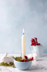 Fototapeten White burning candle, moss, briar berries and white bunnies on white marble background against blue  textured wall. Rabbit is symbool of 2023.. Selective focus. Place for text. © daffodilred